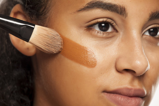 Fototapeta portrait of a young dark-skinned woman applying liquid makeup base with brush on her face on a white background