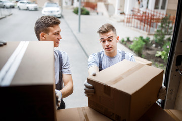 Two young handsome movers wearing uniforms are unloading the van full of boxes. House move, mover service.