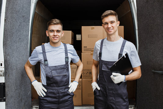Two young handsome smiling workers wearing uniforms are standing in front of the van full of boxes. House move, mover service.