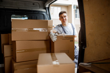 A young handsome smiling worker wearing uniform is standing next to the van full of boxes. House move, mover service.