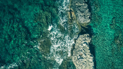 Top down aerial view of the shallow turquoise transparent water with rocks. Sea waves hitting rocks creating foam