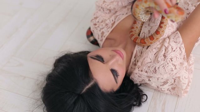 Sexy girl with professional makeup and hairstyle lies on the white wooden floor and holds two snakes, close-up.
