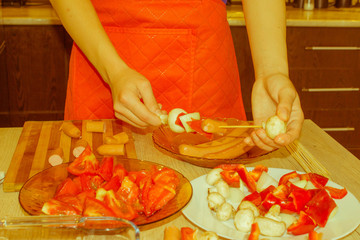 Preparing shashlik for barbecue grill. A hand puts pieces of mushrooms, sausages, tomatoes, pepper...