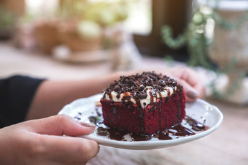 A woman holding a piece of red velvet cake with cream and chocolate in white ceramic plate on wooden table