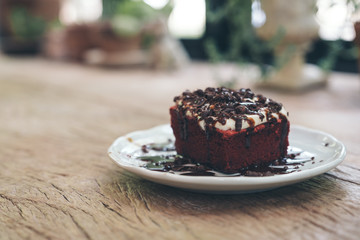 A piece of red velvet cake with cream and chocolate in white ceramic plate on wooden table