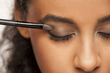 portrait of a young dark-skinned woman applying eye shadow with brush on a white background - obrazy, fototapety, plakaty