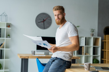 Bearded young businessman working at modern office.Man wearing white t-shirt and making notes on the documents.