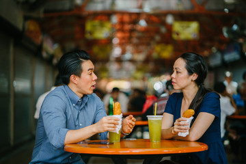 Image of  a good looking Chinese Asian young couple enjoying a snack at a hawker center Singapore, Asia. They are smiling as they converse and enjoy their sugarcane juice and fried bananas. 