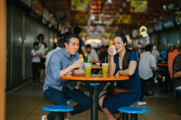 An image of a loving Chinese couple eating their snacks together. They are both smiling and enjoying while having their date at a hawker center..
