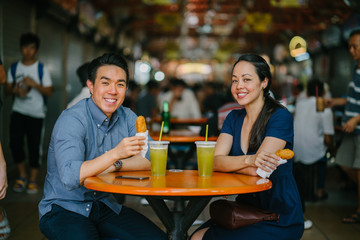An image of a young couple eating together during their date at a hawker center.  They are enjoying their fried banana fritters and sugarcane juice.