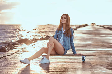 Sensual brunette girl dressed in shorts and a t-shirt is sitting on a skateboard on the beach...
