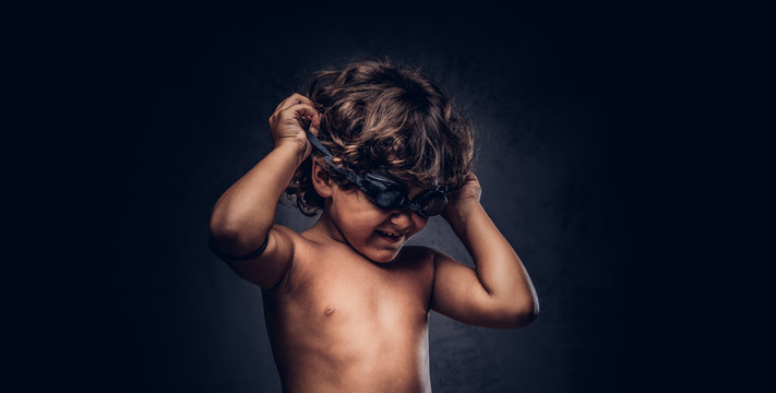 Cute little shirtless boy puts on swimming goggles posing in a studio. Isolated on the dark textured background.
