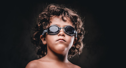 Cute little shirtless boy in swimming goggles posing in a studio. Isolated on the dark textured background.