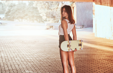 Back view of a sensual young skater girl dressed in shorts and t-shirt holds skateboard and looking at the camera under the bridge. © Fxquadro