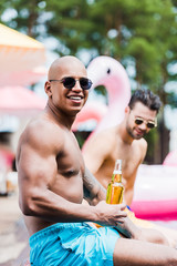 smiling muscular man with beer looking at camera while his friend sitting behind on poolside