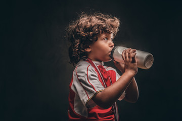 Fototapeta na wymiar Little champion boy in sportswear with a gold medal drinking water from a bottle. Isolated on a dark textured background.