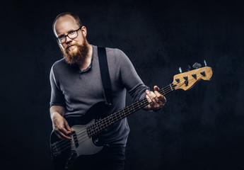 Redhead bearded male musician wearing glasses dressed in a gray t-shirt playing on electric guitar. Isolated on dark textured background.