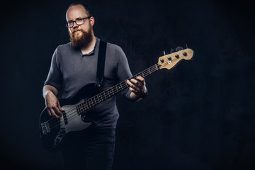 Redhead bearded male musician wearing glasses dressed in a gray t-shirt playing on electric guitar. Isolated on dark textured background.