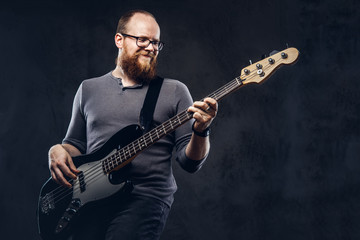 Redhead bearded male musician wearing glasses dressed in a gray t-shirt enjoying playing on electric guitar. Isolated on dark textured background.