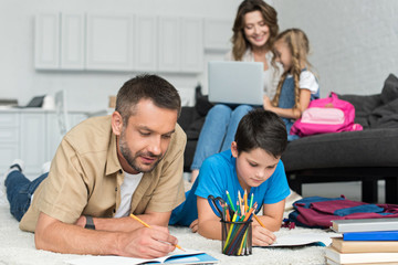 selective focus of father and son doing homework together while mother and daughter using laptop on sofa at home