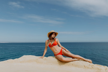Young woman in straw hat and red bikini lying on the stone edge with beautiful view on ocean. Summer vocation