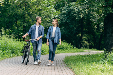beautiful young couple walking by park with vintage bicycle and looking at each other