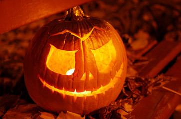 Halloween pumpkin with candle lights  flame sitting on a bench with autumn leaves in a city park at...