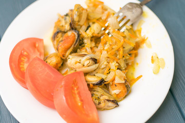Fried mussels with onions, carrots and spices on a white plate with a sliced tomato.