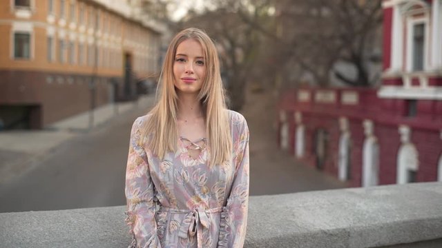 Portrait of cute blonde woman 20s with blond hair wearing dress smiling, while standing on bridge in downtown against road traffic slow motion