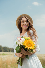 portrait of cheerful woman in white dress with bouquet of wild flowers in field