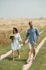 Naklejka na ściany i meble woman in white dress with bouquet of wild flowers walking together with boyfriend in field
