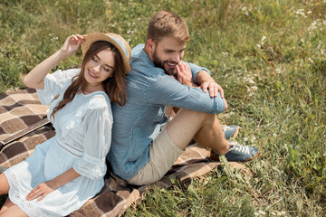 high angle view of smiling couple resting back to back on blanket in field with wild flowers on summer day