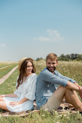 Fototapeta premium young couple resting back to back on blanket in field with wild flowers on summer day