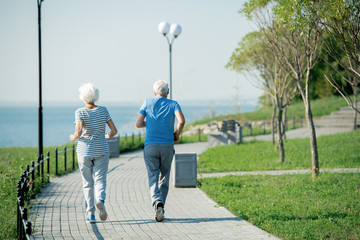 Back view portrait of active senior couple running on park lane along sea shore outdoors , copy...