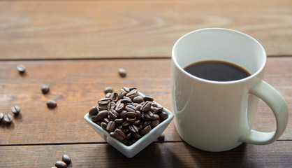 A cup of coffee with small white ceramic dish full of coffee beans on wooden background.