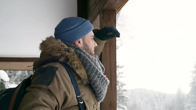 Male tourist with backpack standing in wooden shelter and looking at winter forest while taking a break during hiking in national park at snowy day