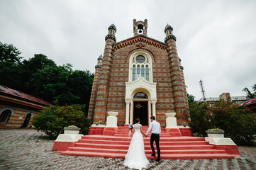Bride dressed in white dress and groom in suit going together on red carpet. Newlyweds going to church before wedding ceremony.