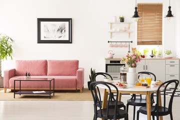 Dining table with breakfast and fresh flowers standing in bright apartment interior with pink sofa,...