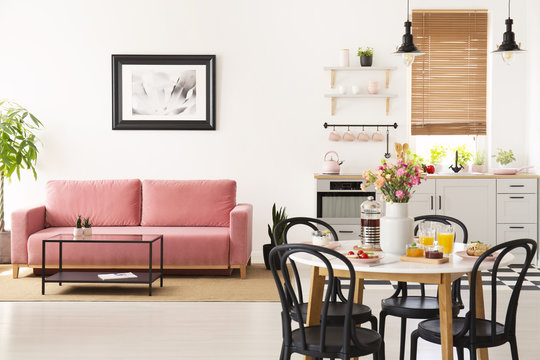 Dining table with breakfast and fresh flowers standing in bright apartment interior with pink sofa, black and white poster and kichenette with pastel accessories