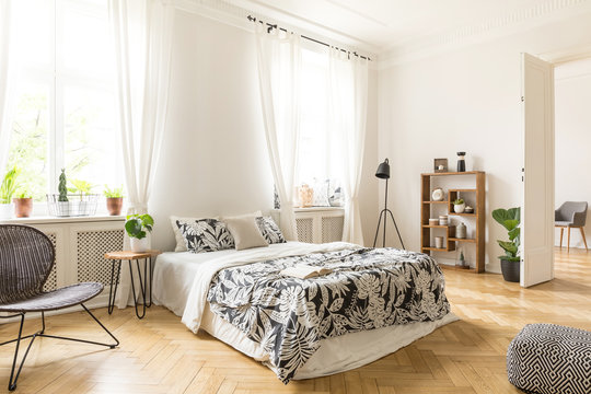 An interior of a bedroom with herringbone parquet, white walls and simple furniture. Bed, chair, bookcase, lamp, stool and pouf inside. Real photo.