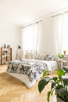 High ceiling bedroom interior with a comfortable bed with leaf pattern linen, a lamp and a bookcase. Real photo.