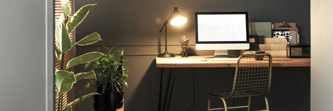 Creative golden, metal chair by a wooden desk with computer screen, folders and an industrial luminous lamp in a stylish home office interior