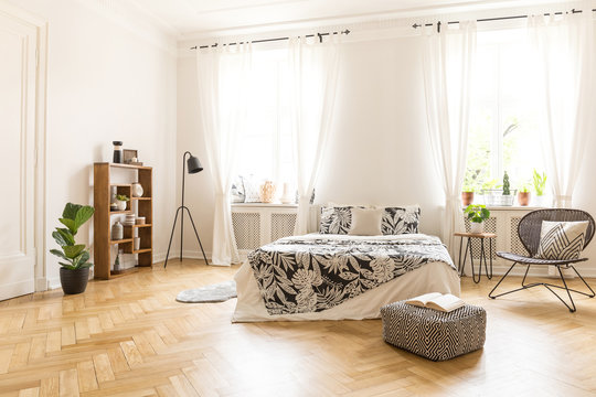 A big bed with linen, a metal and rattan chair, a lamp, a pouf and a bookcase standing on a wooden floor in a spacious bedroom interior with white walls. Real photo.