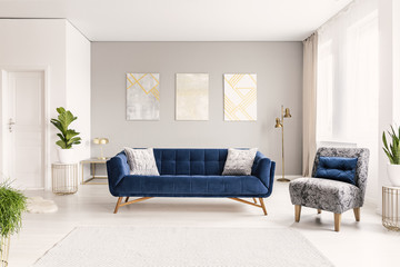 Real photo of a spacious living room interior with a blue sofa, armchair and three paintings
