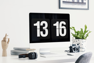 Computer screen showing time, headphones, camera, books, wooden hand an plant on a desk in a bright...