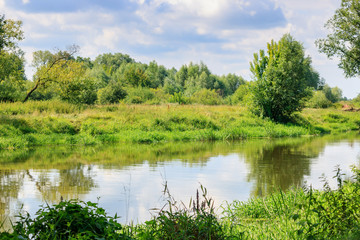 Small river on the background of grass-covered banks against cloudy sky at sunny summer day. River landscape