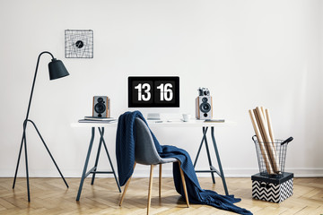 Real photo of a home office interior with a desk, chair with a blanket, lamp, computer and basket...