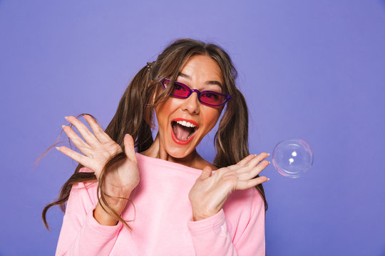 Portrait of glamour attractive woman with two ponytails in sweatshirt wearing trendy sunglasses screaming in delight, isolated over violet background in studio