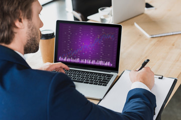 cropped shot of businessman taking notes on clipboard and using laptop with business charts on screen