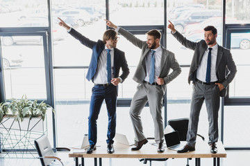 three happy young businessmen dancing on table in office